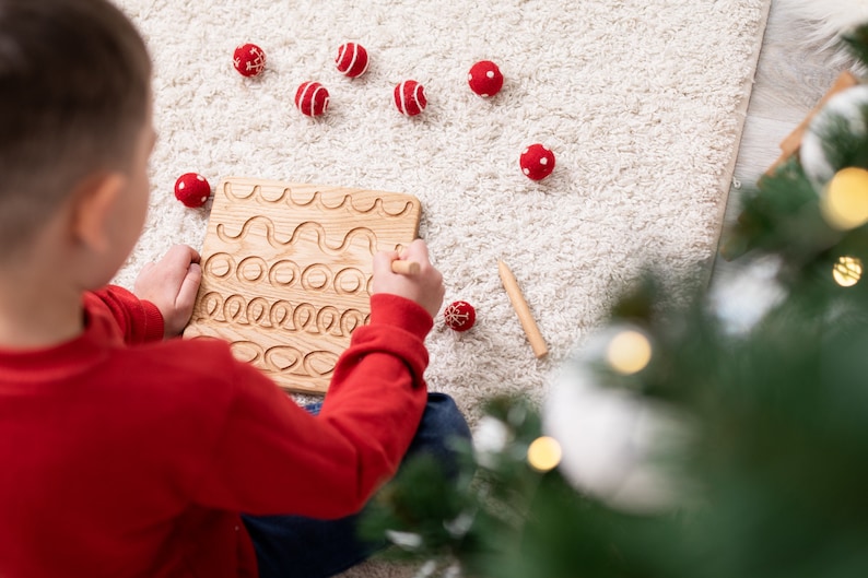May include: A wooden tracing board with carved lines and circles, and a wooden stylus. Scattered around are red and white felt balls. The scene is set on a white rug, with a Christmas tree in the background.