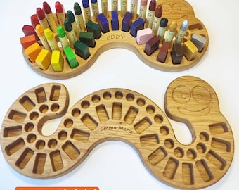 May include: Two wooden crayon organisers, one filled with colourful crayons. The organisers are worm-shaped with a smiling face on the end. One organiser is labelled "Eddy", the other "Emma Marie". The text "crayons not included" is also visible.