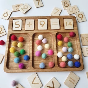 May include: Wooden educational toy with a tray, wooden number tiles, and colorful felt balls. The tray has sections for numbers and math symbols. The numbers 5, 9, and 14 are displayed, along with the plus and equals signs. Various colored balls are in the tray.