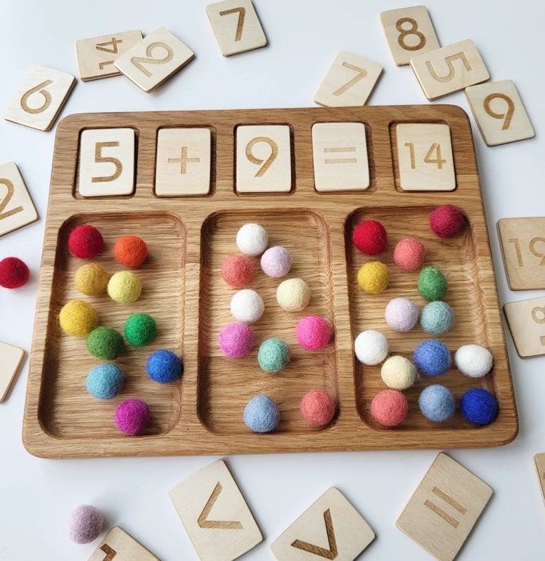 May include: Wooden educational toy with a tray, wooden number tiles, and colorful felt balls. The tray has sections for numbers and math symbols. The numbers 5, 9, and 14 are displayed, along with the plus and equals signs. Various colored balls are in the tray.