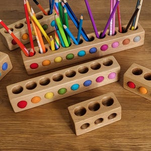 Puede incluir: Una colecci&oacute;n de portal&aacute;pices rectangulares de madera con aberturas circulares e inserciones de fieltro de colores. Los soportes var&iacute;an en longitud y est&aacute;n dispuestos con varios l&aacute;pices de colores colocados en los orificios del bloque m&aacute;s grande.