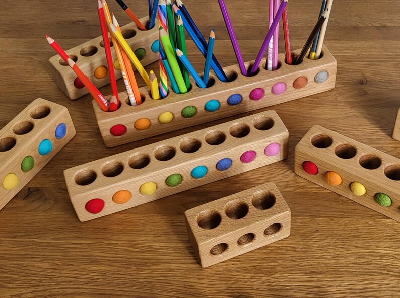 Puede incluir: Una colecci&oacute;n de portal&aacute;pices rectangulares de madera con aberturas circulares e inserciones de fieltro de colores. Los soportes var&iacute;an en longitud y est&aacute;n dispuestos con varios l&aacute;pices de colores colocados en los orificios del bloque m&aacute;s grande.