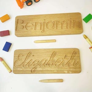 May include: Two rectangular light brown wooden boards featuring carved names. The top board has the name Benjamin in a block font, and the bottom board has the name Elizabeth in a cursive script. Each board includes a matching wooden stylus.