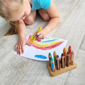 May include: A child drawing a rainbow with a yellow crayon on white paper. A wooden pencil holder with colorful pencils is in the foreground. The drawing includes the word "RAINBOW".