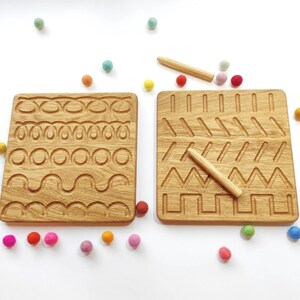 May include: Two wooden tracing boards with grooves of various shapes and patterns. Each board has a wooden stylus. Colorful felt balls are scattered around the boards on a white surface. Educational toy.