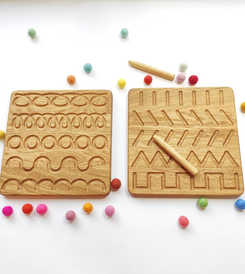 May include: Two wooden tracing boards with grooves of various shapes and patterns. Each board has a wooden stylus. Colorful felt balls are scattered around the boards on a white surface. Educational toy.