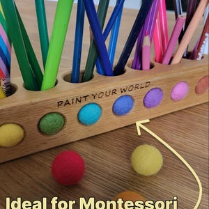 Puede incluir: Un portal&aacute;pices de madera clara con una fila de orificios circulares que contienen varios l&aacute;pices de colores. El frente presenta el texto grabado PAINT YOUR WORLD y una fila de bolas de fieltro de colores. Hay m&aacute;s bolas frente a &eacute;l.