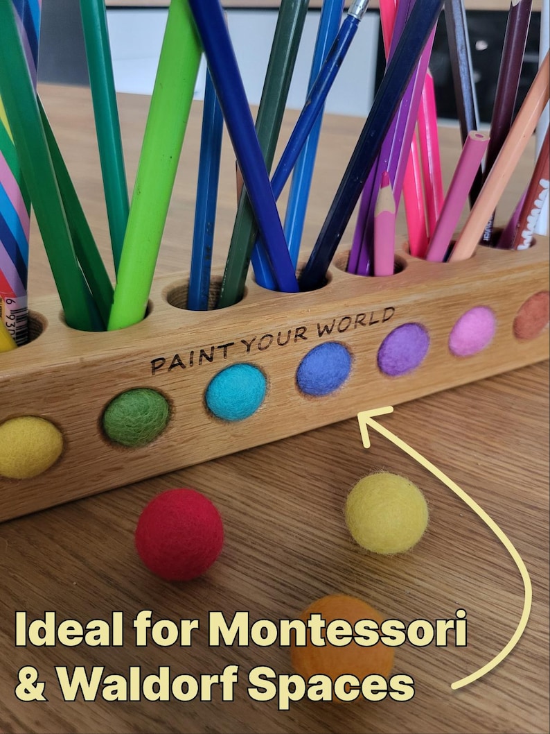 Puede incluir: Un portal&aacute;pices de madera clara con una fila de orificios circulares que contienen varios l&aacute;pices de colores. El frente presenta el texto grabado PAINT YOUR WORLD y una fila de bolas de fieltro de colores. Hay m&aacute;s bolas frente a &eacute;l.
