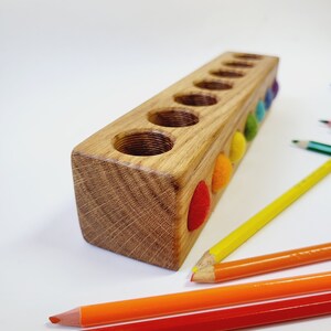 Puede incluir: Un bloque de madera rectangular con siete orificios circulares perforados en la parte superior. El lateral del bloque presenta siete peque&ntilde;as bolas de fieltro de colores en rojo, naranja, amarillo, verde, azul y morado. Cuatro l&aacute;pices de colores.