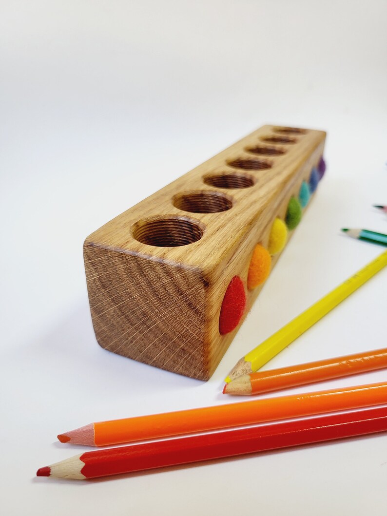 Puede incluir: Un bloque de madera rectangular con siete orificios circulares perforados en la parte superior. El lateral del bloque presenta siete peque&ntilde;as bolas de fieltro de colores en rojo, naranja, amarillo, verde, azul y morado. Cuatro l&aacute;pices de colores.
