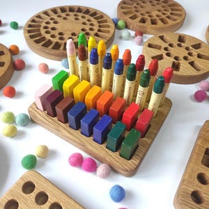 Könnte beinhalten: Halterungen für Buntstifte und Wachsmalblöcke aus Holz mit farbigen Buntstiften und Filzkugeln. Die Halterungen sind aus hellem Holz gefertigt. Der Text "crayons not included" ist im Bild zu sehen.