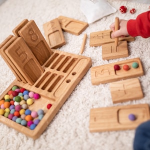 Könnte beinhalten: Holzsatz zum Lernen von Zahlen mit bunten Kugeln, einem Tablett mit Schlitzen für die Zahlen und einem Stift zum Nachzeichnen der Zahlen. Das Spielzeug befindet sich auf einem weißen Teppich.