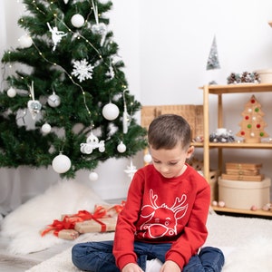 May include: A young boy wearing a red sweater with a white reindeer design sits on a white rug in front of a Christmas tree. He is playing with a wooden toy with metal screws and nuts.