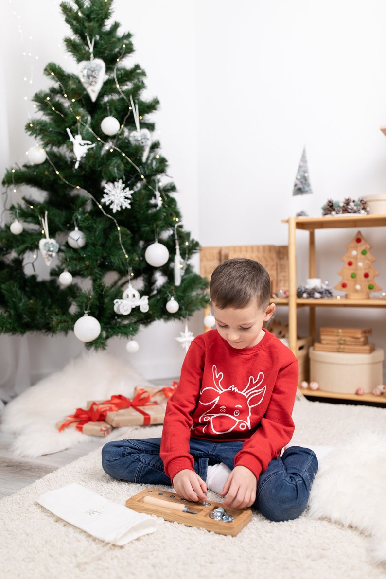 May include: A young boy wearing a red sweater with a white reindeer design sits on a white rug in front of a Christmas tree. He is playing with a wooden toy with metal screws and nuts.
