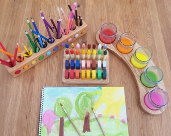 May include: A wooden tray with five small glass jars filled with colourful paint. A wooden tray with 24 small blocks of coloured paint. A wooden pencil holder with 12 coloured pencils. A sketchbook with a watercolour painting of trees and a sun.