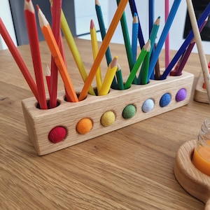 Puede incluir: Un portal&aacute;pices rectangular de madera con m&uacute;ltiples orificios verticales que contienen l&aacute;pices de colores variados. La parte frontal del bloque de madera clara presenta una hilera de peque&ntilde;as bolas de lana fieltrada de colores.