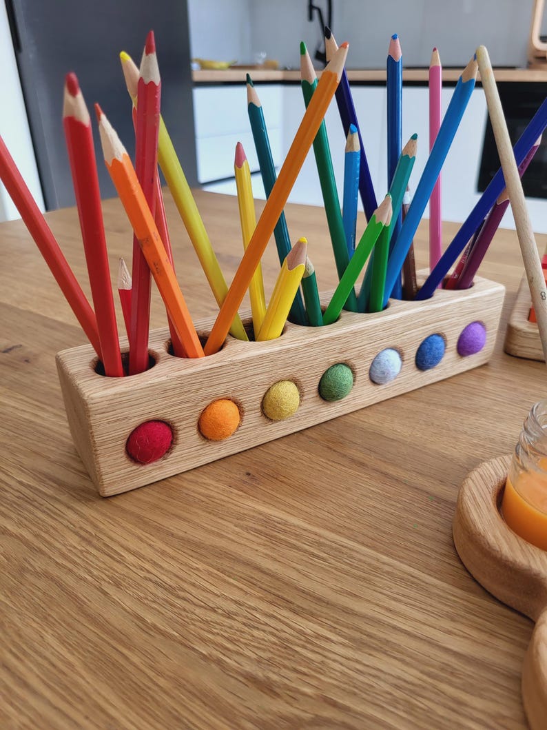 Puede incluir: Un portal&aacute;pices rectangular de madera con m&uacute;ltiples orificios verticales que contienen l&aacute;pices de colores variados. La parte frontal del bloque de madera clara presenta una hilera de peque&ntilde;as bolas de lana fieltrada de colores.