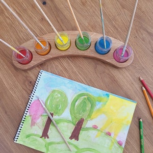 May include: A wooden paint palette with six small jars filled with red, orange, yellow, green, blue, and pink paint. Paintbrushes are in the jars. A watercolour painting of trees and a sun is next to the palette. Coloured pencils are nearby.