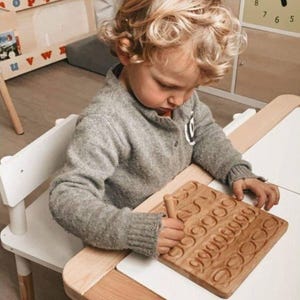 May include: A child is using a wooden tracing board with carved numbers. The board is light brown and sits on a white surface. The child is wearing a gray sweater and is seated at a small white table.
