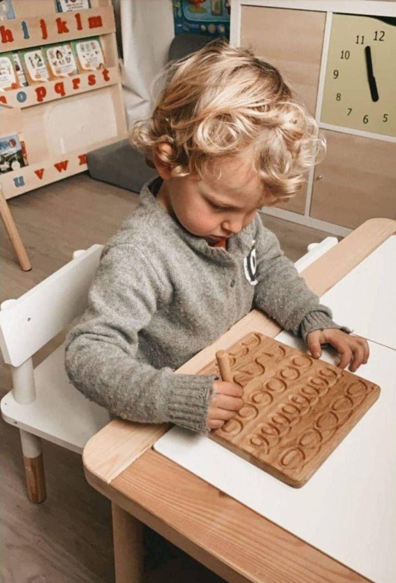 May include: A child is using a wooden tracing board with carved numbers. The board is light brown and sits on a white surface. The child is wearing a gray sweater and is seated at a small white table.