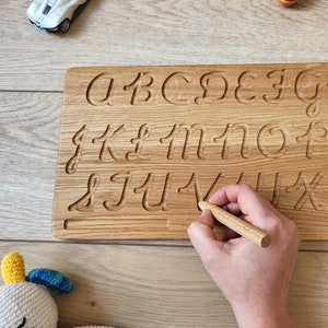 May include: A wooden alphabet tracing board with carved lowercase letters. A small wooden stylus is held in a hand, tracing the letter "V". Toy cars and a spinning top are in the background. A crocheted stuffed animal is in the lower left corner.