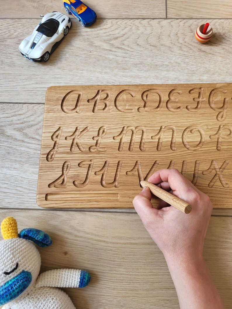May include: A wooden alphabet tracing board with carved lowercase letters. A small wooden stylus is held in a hand, tracing the letter "V". Toy cars and a spinning top are in the background. A crocheted stuffed animal is in the lower left corner.