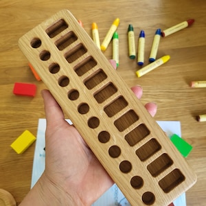 May include: A light brown wooden crayon holder with rectangular and circular cutouts. It holds several colourful crayons. Various crayons and small wooden blocks are scattered around the holder.