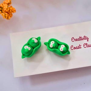 Könnte beinhalten: Ein Paar grüne Erbsenhülsen-Ohrringe mit weißen Gesichtern. Die Ohrringe befinden sich auf einer weißen Karte mit dem Text "Creativity Coast Clay".