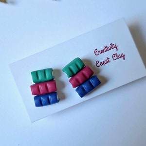 Peut inclure: Une paire de boucles d'oreilles en pâte polymère faites à la main, avec une pile de trois blocs colorés. Les blocs sont verts, rouges et bleus. Les boucles d'oreilles sont présentées sur une carte blanche avec le texte "Creativity Coast Clay".