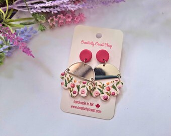 May include: A pair of pink and white floral dangle earrings with silver accents. The earrings are handmade in NB and feature a floral pattern with pink and white flowers. The earrings are displayed on a white card with the text "Creativity Coast Clay" and "handmade in NB www.creativitycoast.com".