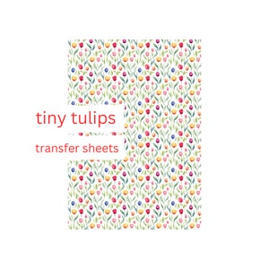 Puede incluir: Una hoja de papel de transferencia con un patrón repetitivo de pequeñas tulipanes de acuarela en tonos de rosa, rojo, amarillo, azul y verde. El texto "tiny tulips transfer sheets" está impreso en la hoja.