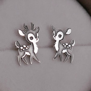 Puede incluir: Un par de pendientes de botón plateados con forma de ciervo. Cada pendiente presenta un diseño detallado de ciervo con cuernos, manchas y una expresión dulce. Los pendientes se exhiben en una caja de joyería de madera con interior gris suave.