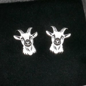 Puede incluir: Un par de pendientes de plata con diseño de cabeza de cabra, presentados en una pequeña caja de regalo. Los pendientes presentan diseños detallados de cabeza de cabra. La caja es gris con un estampado floral.