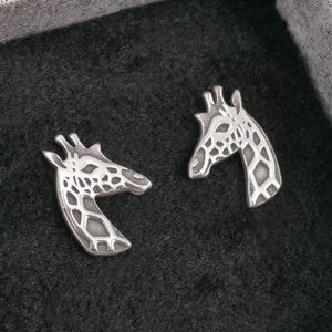 May include: A pair of silver-colored giraffe head stud earrings. The earrings are in a black box with a gray patterned lid. The giraffe heads have a detailed pattern on their faces and necks.