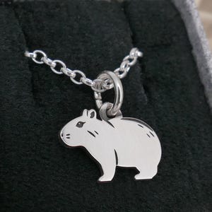 Könnte beinhalten: Eine silberfarbene Halskette mit einem Capybara-Anhänger. Der Anhänger ist eine detaillierte Umrisszeichnung des Tieres mit kleinen geätzten Details. Die Halskette wird in einer schwarzen Schmuckschatulle präsentiert.