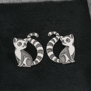 Peut inclure: Une paire de boucles d'oreilles en argent avec un motif de lémurien noir et blanc. Les lémuriens se font face, leurs queues entrelacées.