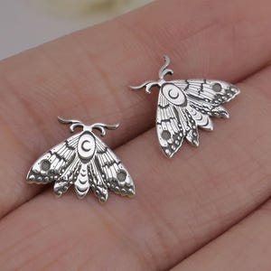May include: A pair of silver-toned earrings featuring a detailed moth design. Each earring has a delicate, intricate pattern of wings and a crescent moon on the body.