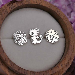May include: Three silver-colored gaming-themed charms: a twenty-sided die, a cartoon dragon, and a six-sided die. The charms are displayed in a wooden ring box with a gray interior, surrounded by purple flowers.