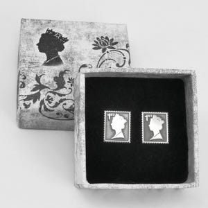 Könnte beinhalten: Eine kleine, weiße Schachtel mit schwarzem Innenraum. Die Schachtel enthält zwei silberne Manschettenknöpfe mit einem Profil eines Frauenkopfes auf jedem. Die Schachtel ist mit einem Blumendesign und einer Silhouette einer Frau verziert.