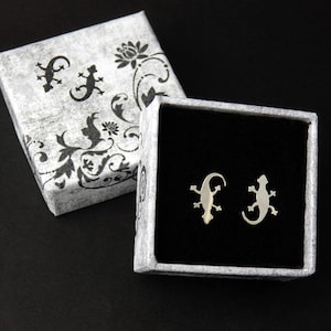 Puede incluir: Una pequeña caja con un patrón blanco y negro con un interior de terciopelo negro. Dos pendientes de lagarto plateados se muestran dentro de la caja.
