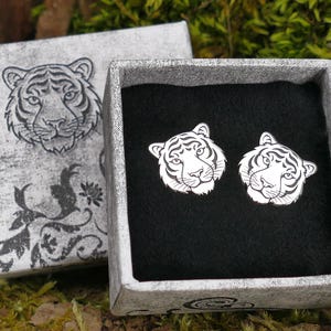 Puede incluir: Un par de pendientes de plata con un diseño de cabeza de tigre blanco, presentado en una pequeña caja gris con un diseño de cabeza de tigre blanco en la tapa.