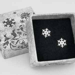 May include: A small, white gift box with a black snowflake design. The box is open and contains two silver snowflake earrings.