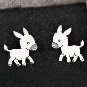 Może przedstawiać: Para srebrnych kolczyków w kształcie osła. Osły patrzą na siebie i mają prosty, kreskówkowy design.