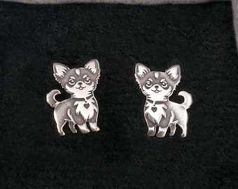 Puede incluir: Un par de broches de perro Chihuahua plateados. Cada broche presenta una representación detallada de un Chihuahua con un colgante en forma de corazón en su collar. Los broches están colocados sobre un fondo negro.