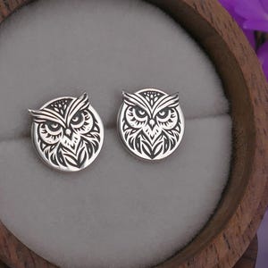 May include: A pair of silver owl stud earrings with a detailed black and white design. The earrings are displayed in a wooden box.