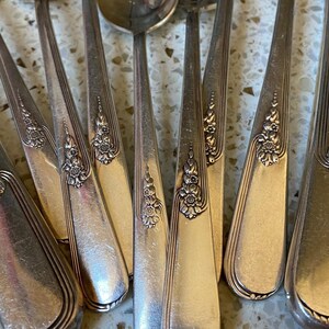 Puede incluir: Un conjunto de cubiertos antiguos, que incluye tenedores, cucharas y un cuchillo. La cubertería plateada presenta detalles florales en los mangos. La disposición se encuentra sobre una superficie clara y moteada.