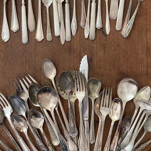 Puede incluir: Un surtido de cubiertos antiguos, que incluye tenedores, cucharas y cuchillos, dispuestos sobre una superficie de madera. La cubertería tiene un acabado plateado y varios diseños de mango. Incluye cucharas de servir y palas para tartas.