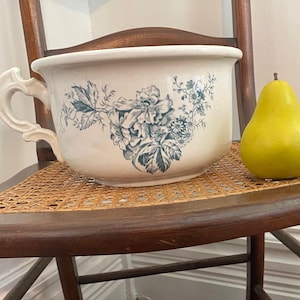 May include: An antique white chamber pot with blue floral designs and a handle sits on a wooden chair with a woven seat. A yellow pear rests beside the pot. The chair is dark wood.