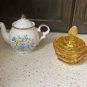 May include: A ceramic teapot with a floral design and a matching lid, next to a small, amber-colored glass jar with a lid. The teapot has a brown handle and spout. The jar has a spiral design.