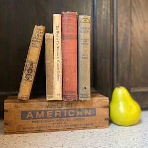 May include: A collection of vintage books, including titles like "The Great Train Robbery" and "American History," displayed on a wooden box marked "American Cheese." A vibrant yellow pear is positioned next to the books, creating a classic aesthetic.